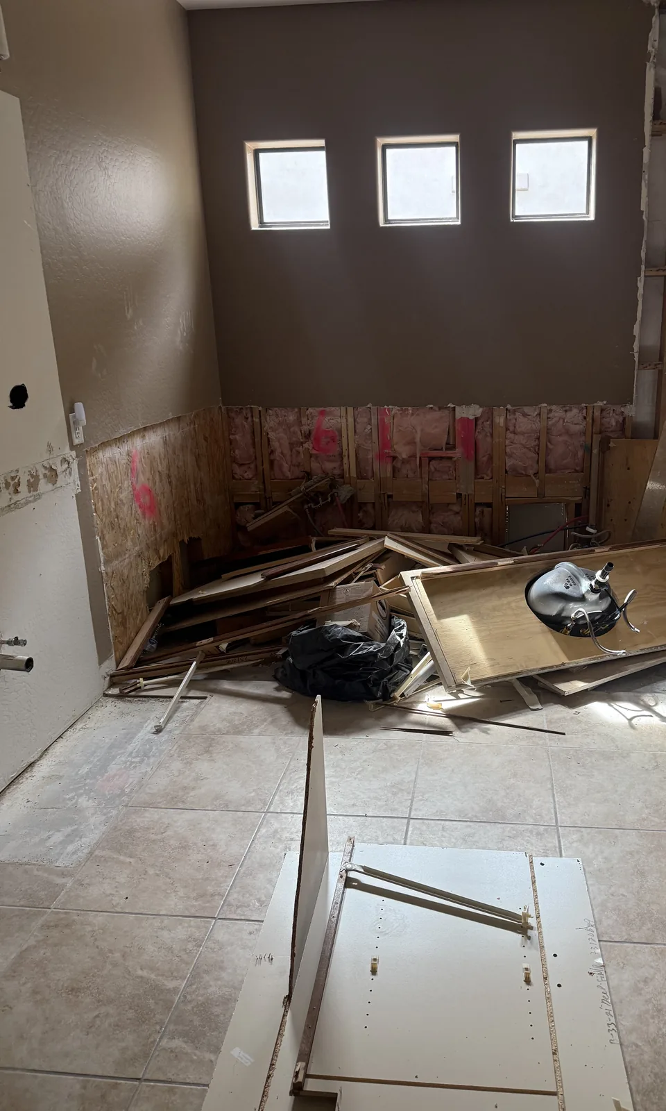 Partly demolished bathroom with exposed studs and insulation, and a pile of broken cabinet boards and scrap on the floor