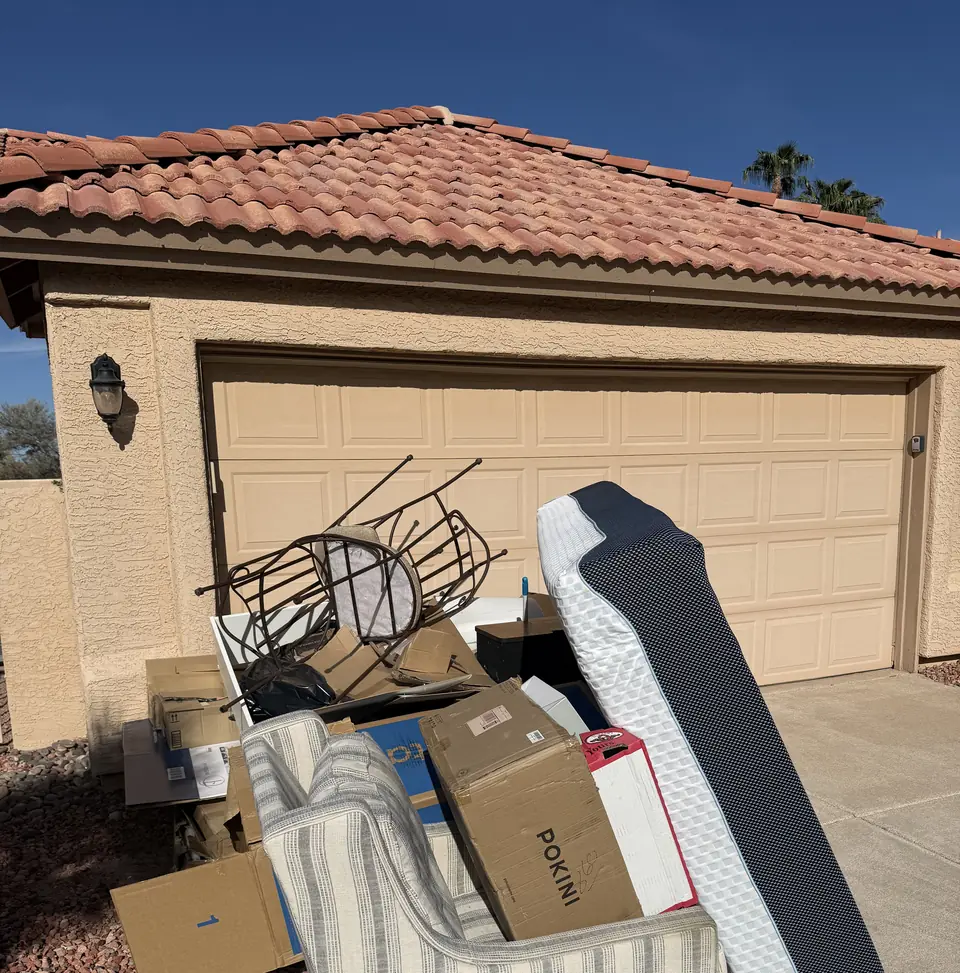 Couch, standing mattress, cardboard boxes and a broken chair piled on a driveway in front of a garage, ready for pickup