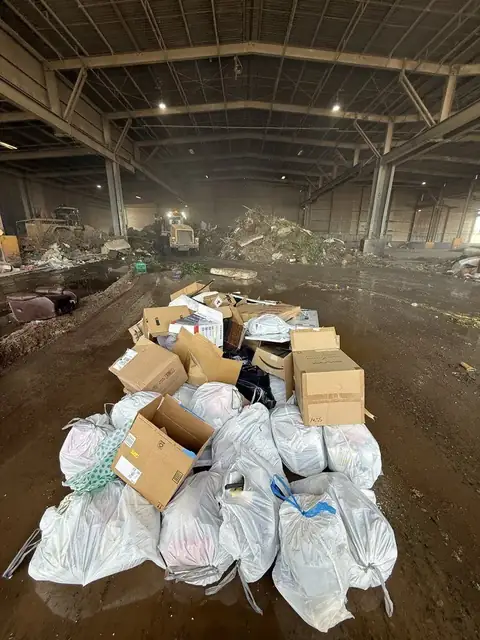 White trash bags and cardboard boxes piled on a dirt floor inside a large waste transfer building