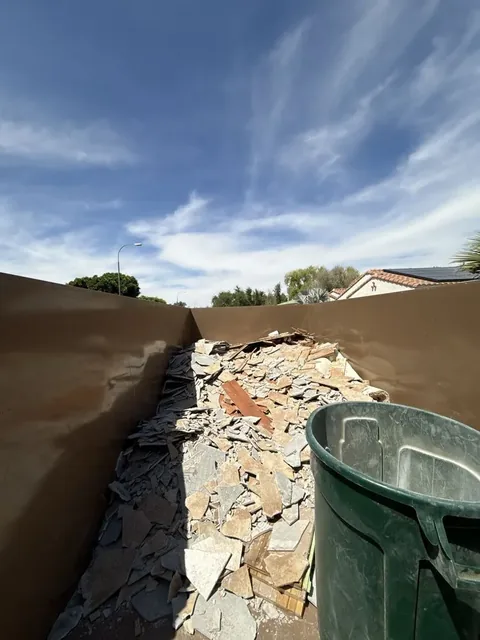 Dump trailer with a heap of broken flagstone and tile, a green trash can beside it and a blue sky overhead