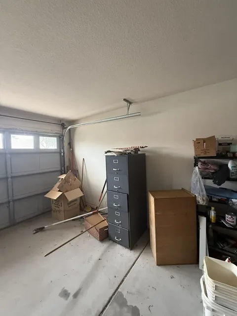 One-car garage with a grey file cabinet, a wooden cabinet, cardboard boxes, brooms and a wire shelf