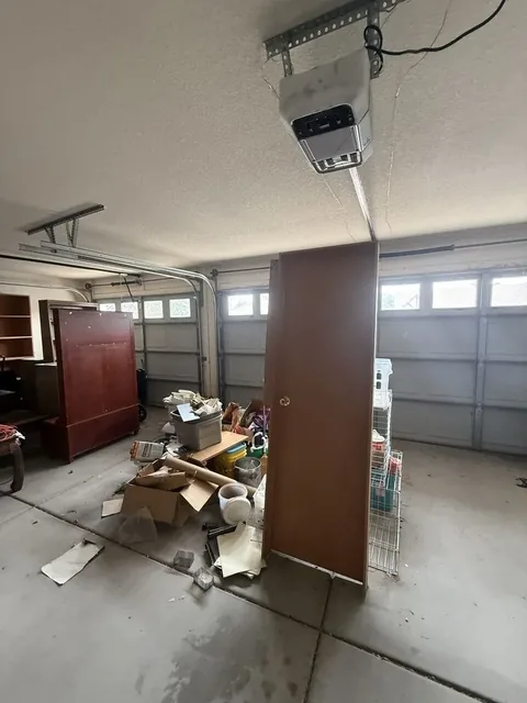 Two-car garage with a tall wooden door on end, a large wooden cabinet, and cardboard and bins piled on the concrete floor