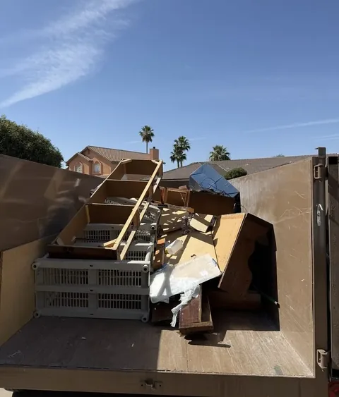 Dump trailer loaded with a wooden shelf unit, plastic crates and boards, with palm trees and a house behind