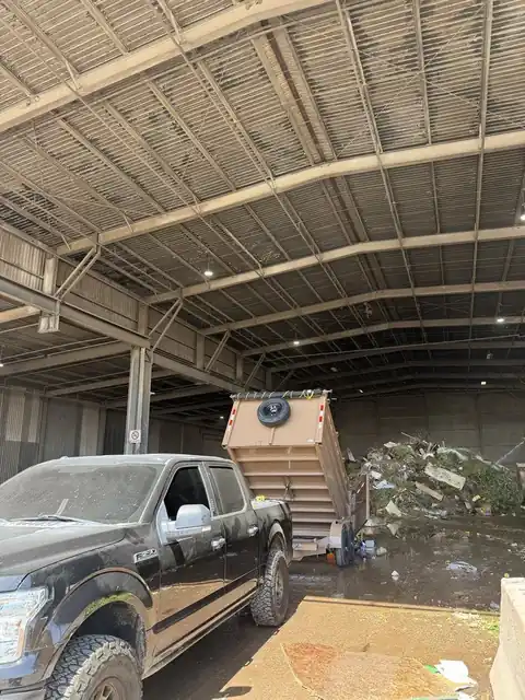 Dark pickup with a tan dump trailer tipped up, unloading at a covered waste transfer station