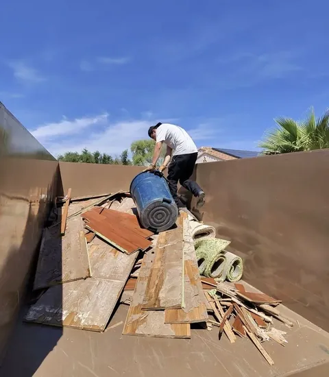 Worker standing in a dump trailer lifting a blue barrel over old floorboards, plywood and rolled green carpet