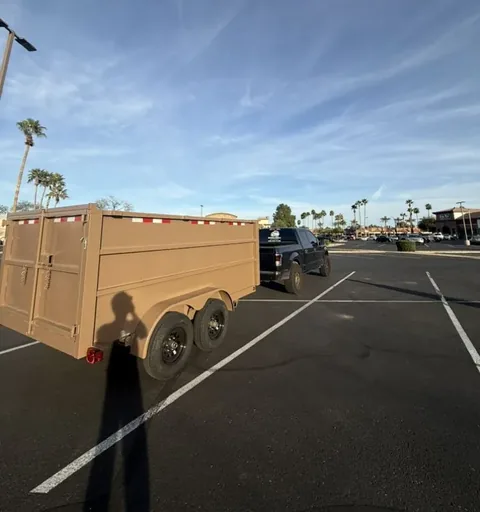 Tan dump trailer hitched to a dark pickup in a shopping-center parking lot with palm trees and a wide sky